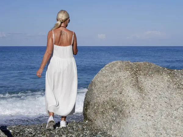 Beyaz elbiseli sarışın kadın güneşli yaz gününde kayalık sahilde akıllı telefonuyla yürüyor.