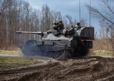 Eğitim tatbikatında çıplak ağaçlar ve bulutlu gökyüzü ile çamurlu arazide manevra yapan PzH 2000NL topçu birliği