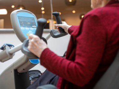Kırmızı kazaklı kişi, kardiyak rehabilitasyon için dijital ekranlı sabit egzersiz bisikleti kullanıyor.