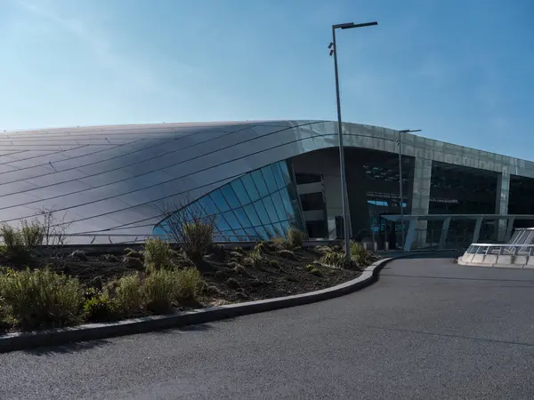 Eğri metalik cepheli modern bina girişi ve mimari tasarımı gösteren peyzaj yaklaşımlı yol