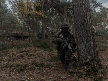 Orman çevresinde savaş teçhizatında askeri personel ve yakınlarda araçlar var.