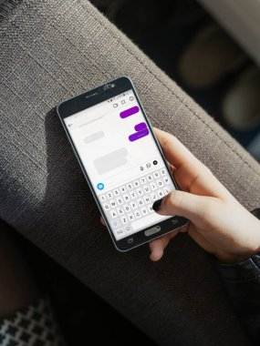 Modern iç mekanda gri döşemeli mobilyalar üzerinde otururken akıllı telefon mesajlaşma uygulaması kullanan kişi