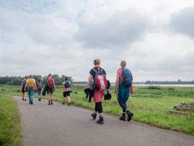 Sırt çantalı çoklu yürüyüşçüler dağınık binalarla yeşil kırsal alanda asfalt yolda yürüyorlar.
