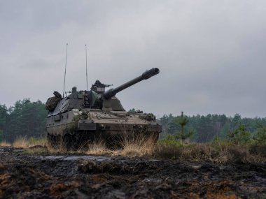 PzH2000NL topçu sisteminin tam görüntüsü orman arka planı ve bulutlu gökyüzü ile engebeli arazide yer almaktadır.