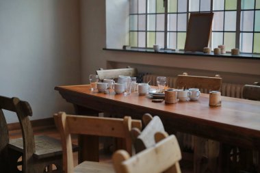 Kırsal ahşap yemek masası, seramik bardaklar ve cam eşyalar geleneksel iç mekanda, vitray pencereli arka planda.