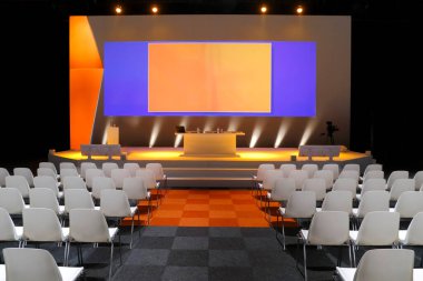 Turuncu sahne grafikleri, beyaz oturma sıraları ve profesyonel AV ekipmanlarıyla boş konferans salonu