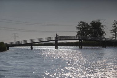 Arka planda elektrik hatları ve ağaçlarla dolu köpüklü suyun üzerinde bisiklet süren insan silueti.