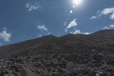 Parlak mavi gökyüzü ve güneşin altında dağınık taşlarla çorak kayalık bir dağ.