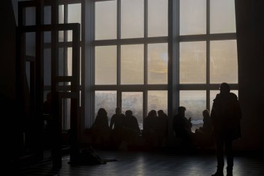 İnsanlar büyük pencerelerde toplanmış altın gün batımını, iç mimari sahneleri izliyorlar.