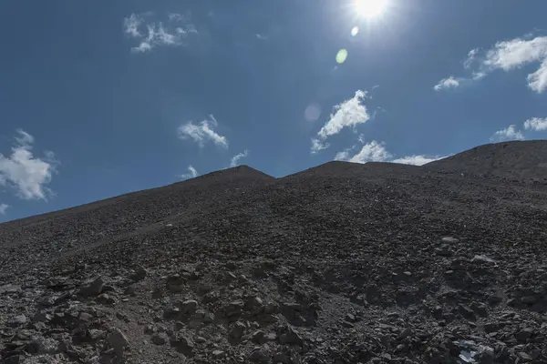 Parlak mavi gökyüzü ve güneşin altında dağınık taşlarla çorak kayalık bir dağ.
