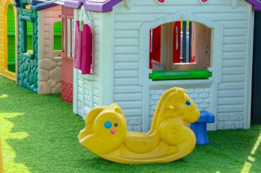 Parlak sarı sallanan at, renkli plastik oyun evlerinin yanında yeşil çimenlerin üzerinde oturur. Bu da çocukların eğlencesi ve eğitici pazarlama için ideal bir açık hava ortamı yaratır..