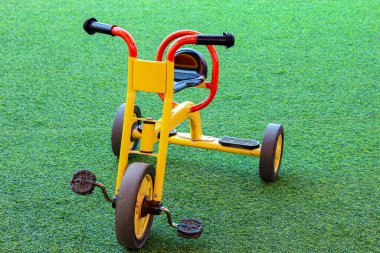 Sarı ve kırmızı üç tekerlekli bisiklet, parlak yeşil suni çimen yüzeyinde sabit durur ve çocuk ürünleri pazarlaması veya aile odaklı tasarımlar için eğlenceli ve davetkar bir sahne yaratır..