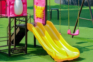 Yeşil bir yüzey üzerinde sarı slayt ve pembe salıncak içeren parlak oyun alanı, çocukların açık hava aktiviteleri için eğlenceli ve davetkar bir alan ve çocukluk ve dinlenme ile ilgili pazarlama için uygun bir ortam sağlıyor..