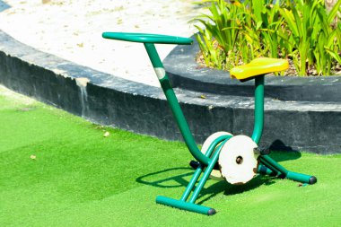 Sarı koltuklu açık yeşil durağan bisiklet park benzeri bir ortamda suni çimlerin üzerinde duruyor. Spor ve eğlence amaçlı yaşam tarzı için güneşli ve davetkar bir atmosfer sağlıyor..