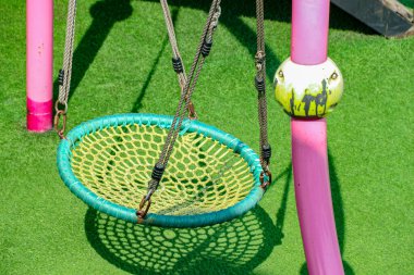 Yuvarlak, net tarzda, yeşil kenarlı ve sarı ağlı bir salıncak, parlak yeşil suni çimenli, çocuk aktiviteleri ve eğlence temaları için ideal, güneşli bir oyun parkındaki pembe direklere bağlanmış iplerden sarkıyor..