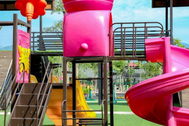 Parlak pembe slayt ve sarı aksanlı bir oyun alanı yapısı, metal destekli yapay yeşil çimenler üzerinde durur, çocukların aktiviteleri veya park temalı tasarımları için açık hava oyunlarının neşeli atmosferini yakalar.