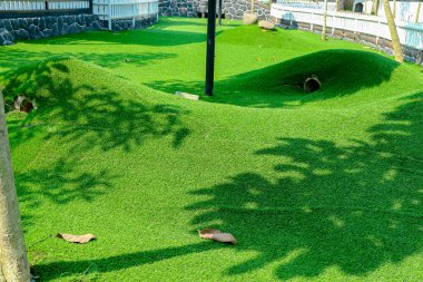 Açık hava manzaralı bir alan oluşturan parlak yeşil suni çimenler bir tünel ile küçük tepe, iç içe geçmiş gölgeler ve dağınık yapraklar, peyzaj ve dış tasarım için ideal.