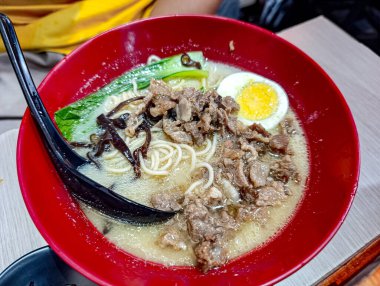 Bir kase iştah açıcı ramen, erişte, sığır eti, bok choy ve yarım haşlanmış yumurtayı kırmızı bir kasede sunar, yemek blogları, restoran promosyonları ve mutfak temalı tasarımlar için davetkar bir görüntü sunar..