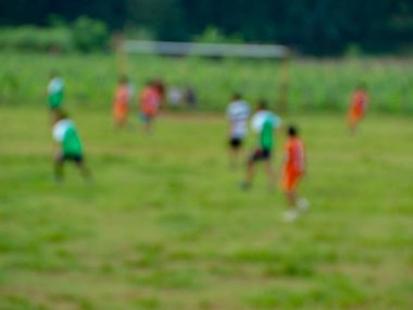 Yeşil çimen tarlasında, uzak ağaçlar ve kale direğiyle futbol oynayan bir grup bulanık çocuk, spor ve eğlence içeriklerine uygun dinamik bir sahne yaratıyor..