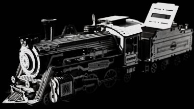 Kontrastlı, siyah ve beyaz bir fotoğraf titizlikle hazırlanmış bir buharlı lokomotifin ahşap modelini ve hassasiyetini yakalar. Lazer kesim parçalardan yapılan model, görünür dişliler, perçinler ve karmaşık mekanik detaylar içeriyor..