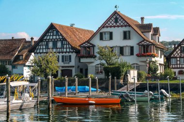 Picturesque lakeside scene featuring traditional European-style houses with steep roofs and timber framing. Small boats, are moored at the dock. Calm water reflects the buildings and greenery, evoking a peaceful village atmosphere.