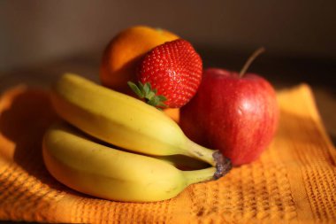 Photograph of fresh fruits arranged on an orange textured cloth. Includes bananas, a red apple, a large strawberry, and an orange. The vibrant colors and close composition highlight the freshness and visual appeal of healthy food choices.