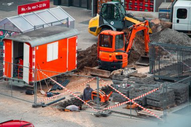 Active construction site in a city area featuring an orange excavator, manual laborer, safety fencing, and mobile office unit. Paving stones, caution tape, and surrounding structures highlight infrastructure development and urban engineering. 