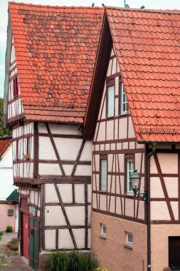 Traditional half-timbered buildings with steep red-tiled roofs and exposed wooden beams in a Central European setting. Moss-covered roof and vintage wall-mounted street lamp add character. Cobblestone path enhance the charm of this old-town scene.
