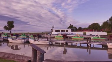 Neusiedler Gölü 'ndeki sakin tekne rıhtımında pastel renkli günbatımında. Küçük tekneler ve iskeleye yanaşmış bir feribot, yumuşak akşam ışığını yansıtıyor..