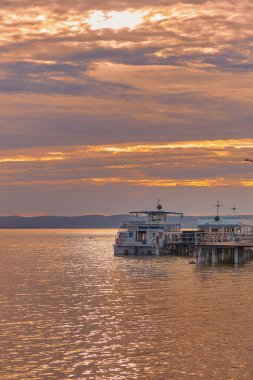 Neusiedl Gölü 'ndeki göl kıyısında dinlenen feribot, sakin bir su ve arka planda parlayan pastel şeftali rengi bir günbatımı gökyüzü..