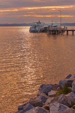Akşam feribotu Avusturya 'nın Neusiedl gölündeki ahşap iskelede, sakin sularla ve huzurlu bir yaz havasıyla pastel şeftali günbatımının altında..