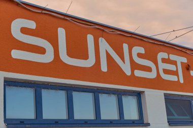 Beyaz günbatımı tabelası ve mavi pencereli turuncu şerit, dramatik ışık ve pastel tonlarıyla sinematik günbatımı gökyüzünün altında parlıyor..