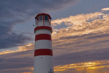 Kırmızı-beyaz Podersdorf Feneri alevli bir turuncu ve pembe gökyüzüne karşı duruyor, günbatımının altın parıltısıyla aydınlanıyor..