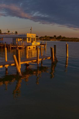 Altın günbatımı ışınları Neusiedler See 'deki ahşap iskeleyi ve yakınlardaki tekneyi aydınlatır. Karanlık yansıtıcı su ve dramatik gökyüzü sinematik bir göl manzarası yaratır..