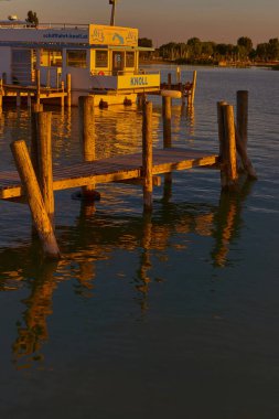 Altın günbatımı ışınları iskeleyi ve demirli tekneyi aydınlatır. Karanlık göl suyu ve dramatik gökyüzü romantik ve sinematik bir Avrupa göl manzarası yaratır..