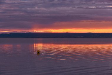 Gün batımında atmosferik dağ gölü, mor bulutlar, suyun üzerinde parlayan turuncu yansıma, sakin yüzeyde sürüklenen barışçıl şamandıra..