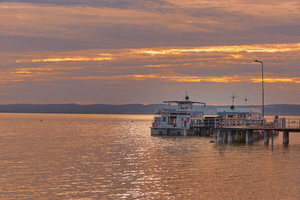Паром пристыковался к деревянному пирсу на озере Нойзидл, Австрия, под ошеломляющим персиковым закатным небом. Спокойные воды и теплый вечерний свет.