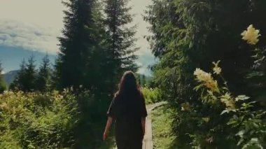 Uzun siyah saçlı bir kız köknar ağaçları arasında tahta bir patika boyunca yürür, çiçeklerle dolu güneşli bir dağlık çayırda ortaya çıkar. Çikolata kıyafeti, Avusturya Alpleri, Styria, sakin dağ manzarası..