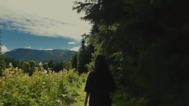 Kız güneşli bir dağ yolunda yürüyor. Solunda çiçek açan alp çiçekleri ve otlar, sağında ise bir sıra köknar ağacı. Yemyeşil Avusturya Alpleri, sakin ve atmosferik dağ manzarası.