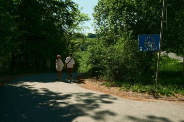 Avusturya 'dan Slovenya' ya geçiş işaretini veren Sdsteirische Weinstrae manzaralı sınır tabelasından geçen bir çift görülüyor. Seyahat, Avrupa keşfi ve kırsal içerik için mükemmel.