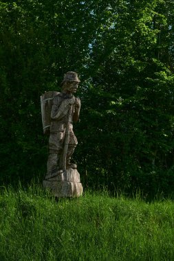 Arka planda yoğun bir Slovenya ormanı bulunan yeşil çimlerin arasında duran ahşap heykel, sakin bir doğal ortamda geleneksel işçiliği temsil ediyor..