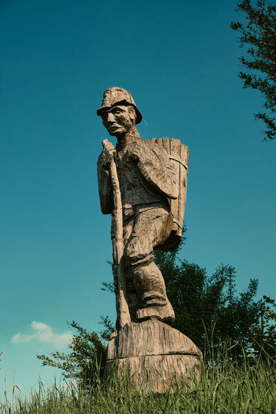 Wooden sculpture of a winemaker with a basket on his back and a staff in hand standing on a hill among green grass and bushes, with a bright blue sky. Symbol of South Styria winemaking traditions
