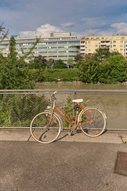 Nehir veya kanal kenarındaki metal korkuluklara park edilmiş turuncu ve beyaz bir bisikletin yakın çekimi, karşı kıyısında yeşil ağaçlar, çalılar ve modern binalar var..