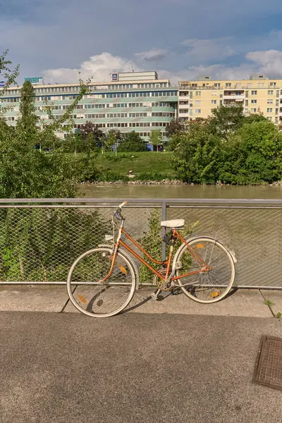 Nehir veya kanal kenarındaki metal korkuluklara park edilmiş turuncu ve beyaz bir bisikletin yakın çekimi, karşı kıyısında yeşil ağaçlar, çalılar ve modern binalar var..