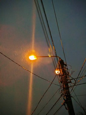 Parlayan turuncu sokak lambaları ve yağan yağmuru aydınlatan elektrik direğinin olduğu dramatik bir akşam sahnesi. Karanlık gökyüzünü kesen ışık demetleri, yağmur damlalarını görünür bir şekilde parlatıyor..