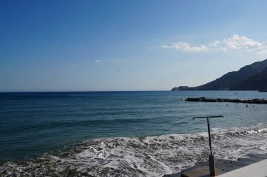 Kıyıda hafif dalgaların dalgalandığı huzurlu sahil manzarası, uzakta yüzücüler, iskelede bir Yunan bayrağı, ve berrak mavi gökyüzünün altında engebeli dağlar..