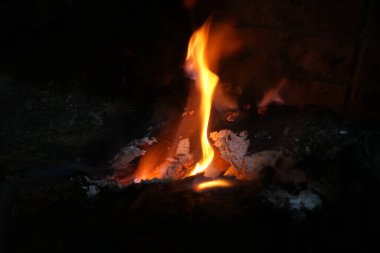 Parlayan közlerden yükselen turuncu bir alevin yakın plan çekimi ve karanlık bir şömine ortamında kömürleşmiş ahşap. Ateşin sıcaklığını, hareketini ve rengini yakalar.