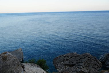 Berrak bir gökyüzünün altındaki kayalık bir uçurumun kenarından sakin mavi deniz manzarası. Minimalist kompozisyon dinginliği, doğayı ve açık alanı vurguluyor..