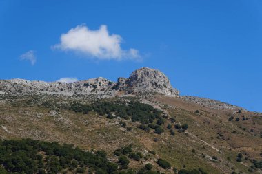 Parlak mavi bulutlu bir gökyüzünün altında bir dağın tepesindeki kireçtaşı oluşumunu vuruyor..