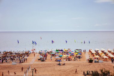 Renkli şemsiyeler, bayraklar ve güneşli bir yaz gününün tadını çıkaran insanlarla dolu canlı bir plaj. Canlı atmosfer, deniz kıyısındaki tatil zamanının zirvesini yakalıyor..